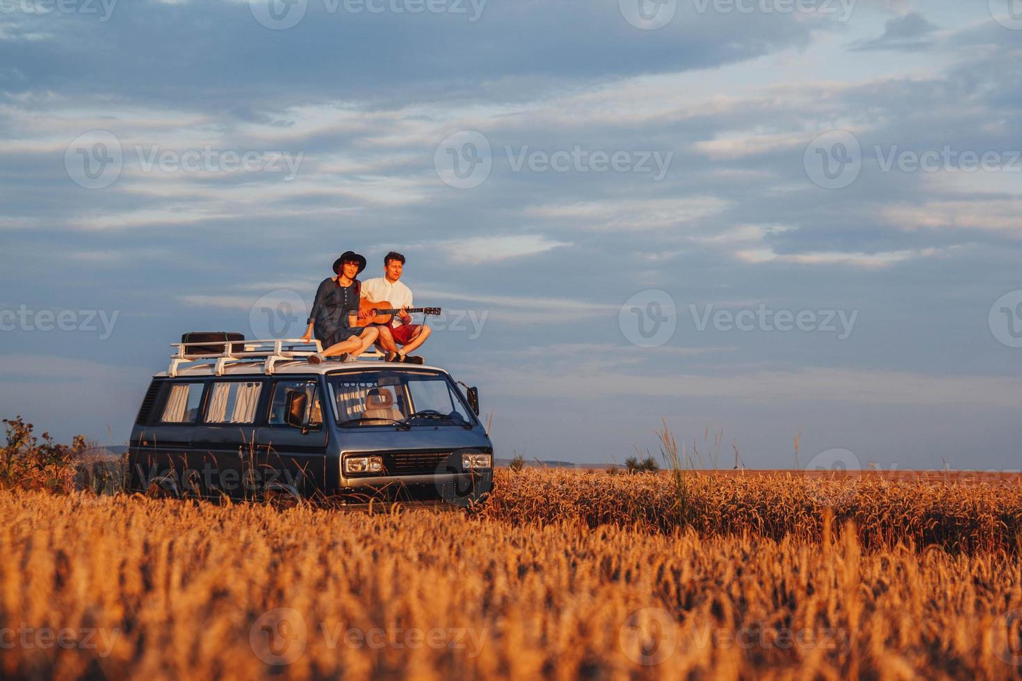 Coppia uomo con una chitarra e una donna con un cappello sono seduti sul tetto di un'auto in un campo di grano foto