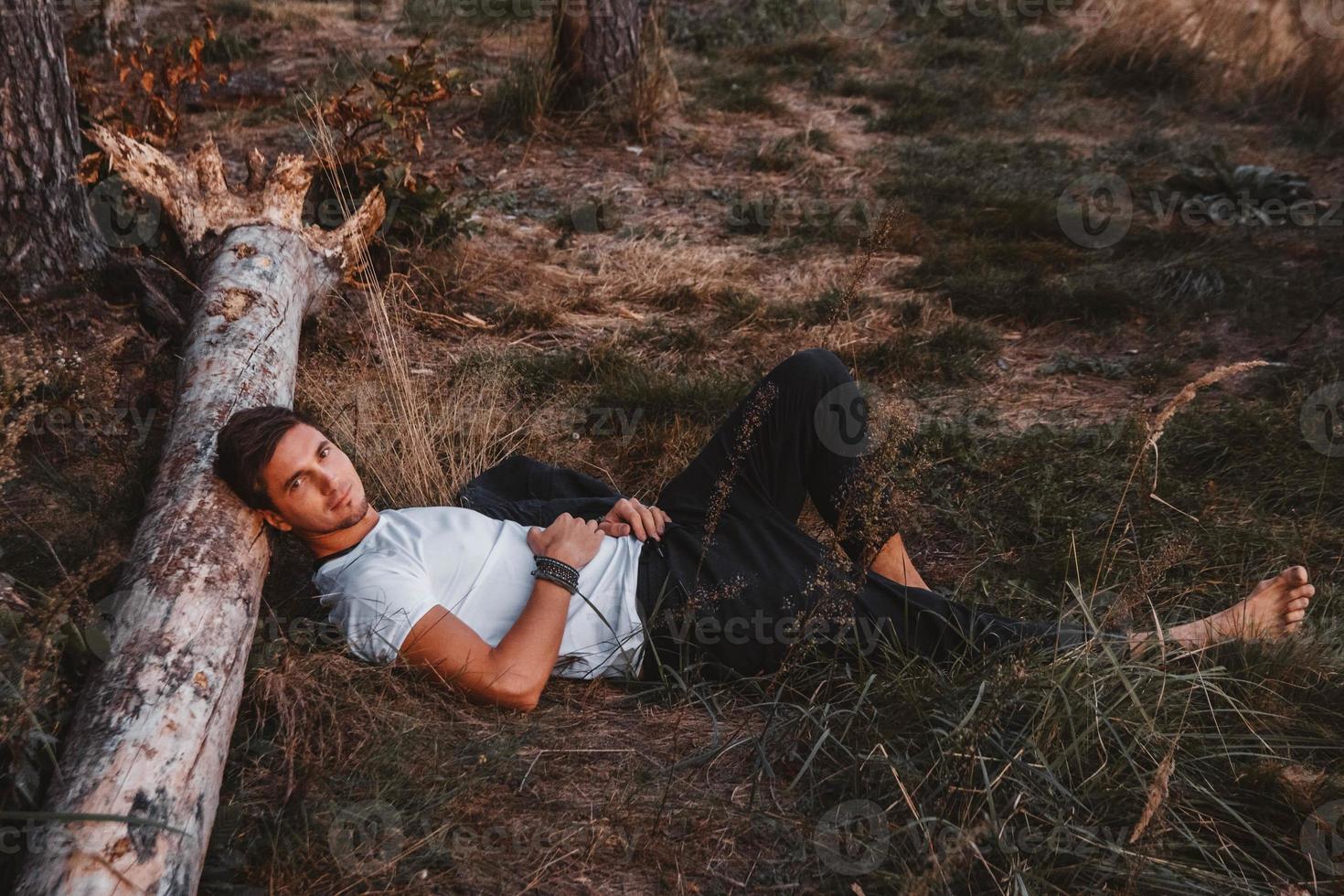 l'uomo giace sull'erba, appoggiando la testa su un tronco in mezzo alla foresta e rilassandosi foto