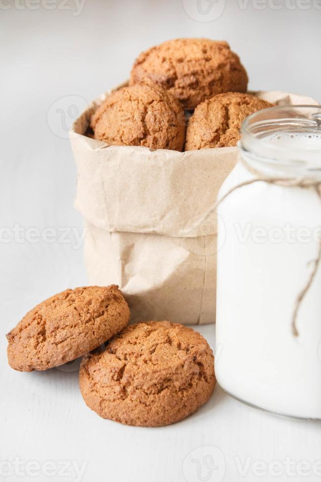 latte in un barattolo di vetro e biscotti di farina d'avena in un sacchetto di carta su un tavolo bianco foto