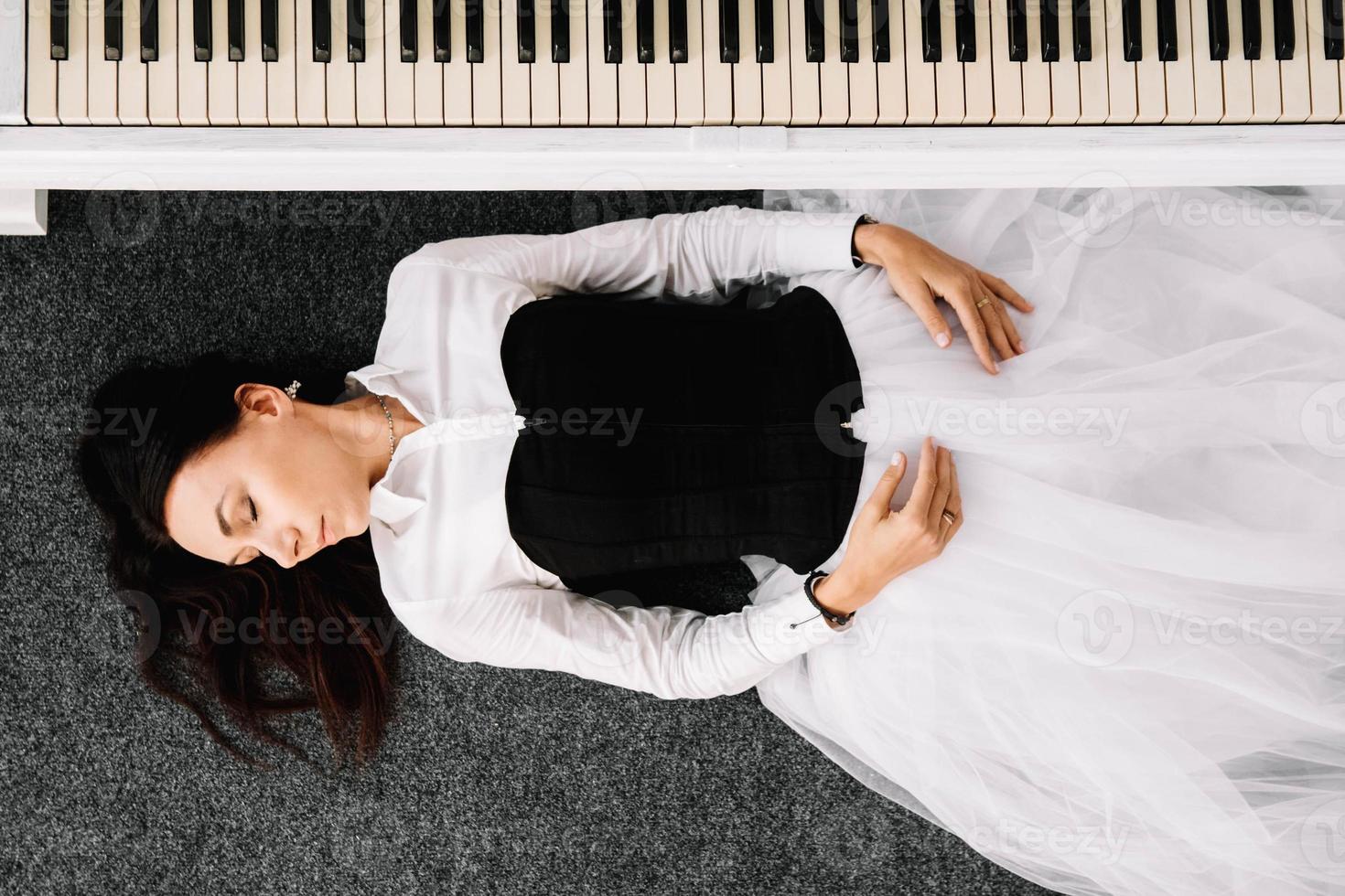 bella donna vestita con un abito bianco con un corsetto nero giace sul pavimento vicino al pianoforte bianco. posto per testo o pubblicità. vista dall'alto foto