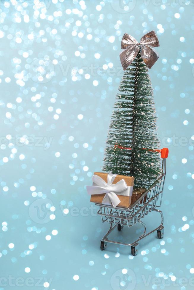 un carrello della spesa con scatole regalo e un albero di natale su uno sfondo colorato con luci. il carrello del supermercato è pieno di regali. concetto di natale di acquisti e vendite online. foto