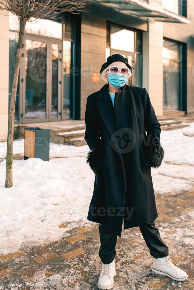 donna alla moda senior in berretto ed elegante cappotto nero e in maschera medica che cammina all'aperto. blocco, pandemia, concetto di protezione foto