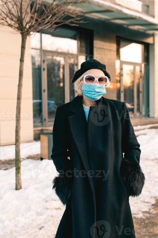 donna alla moda senior in berretto ed elegante cappotto nero e in maschera medica che cammina all'aperto. blocco, pandemia, concetto di protezione foto