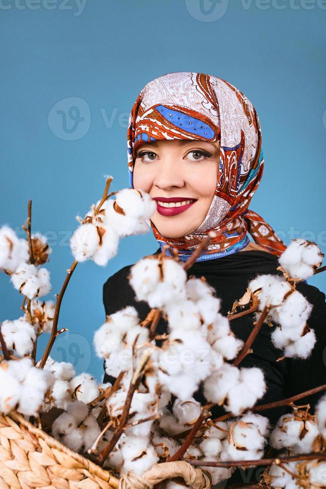 donna orientale in fazzoletto colorato che tiene cesto con rami di cotone su sfondo blu. raccoglitore di cotone foto