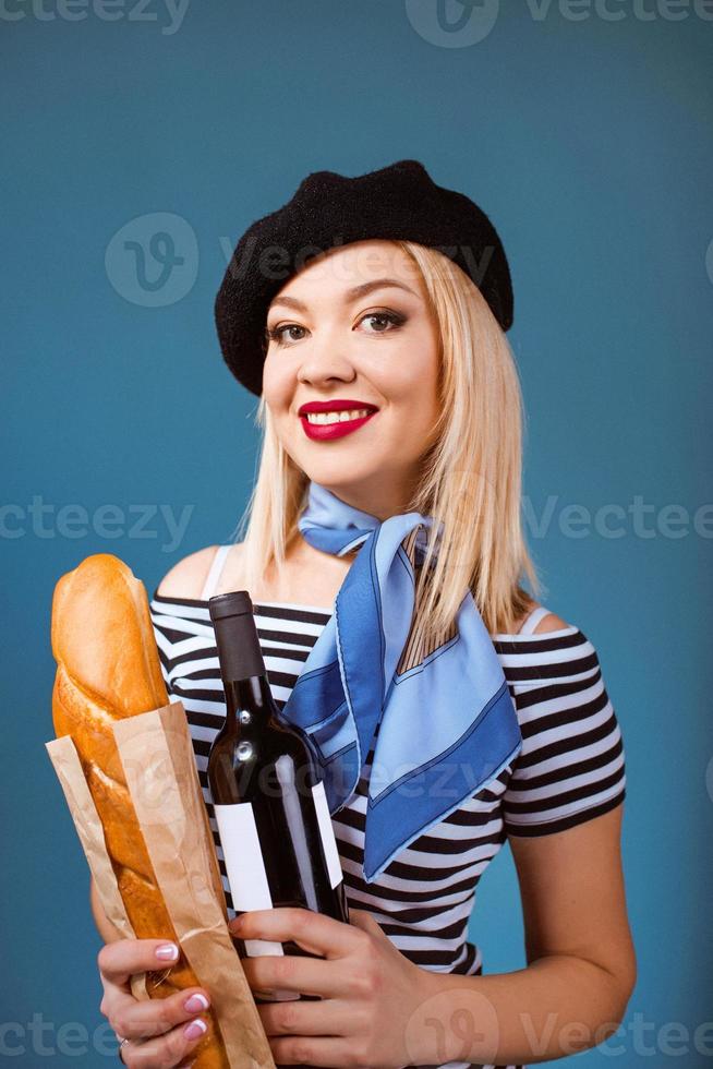 Ritratto di bella bionda donna francese in berretto, sciarpa, schiena e camicia bianca, con bottiglia di vino e baguette di pane in braccio con bandiera francese sullo sfondo foto