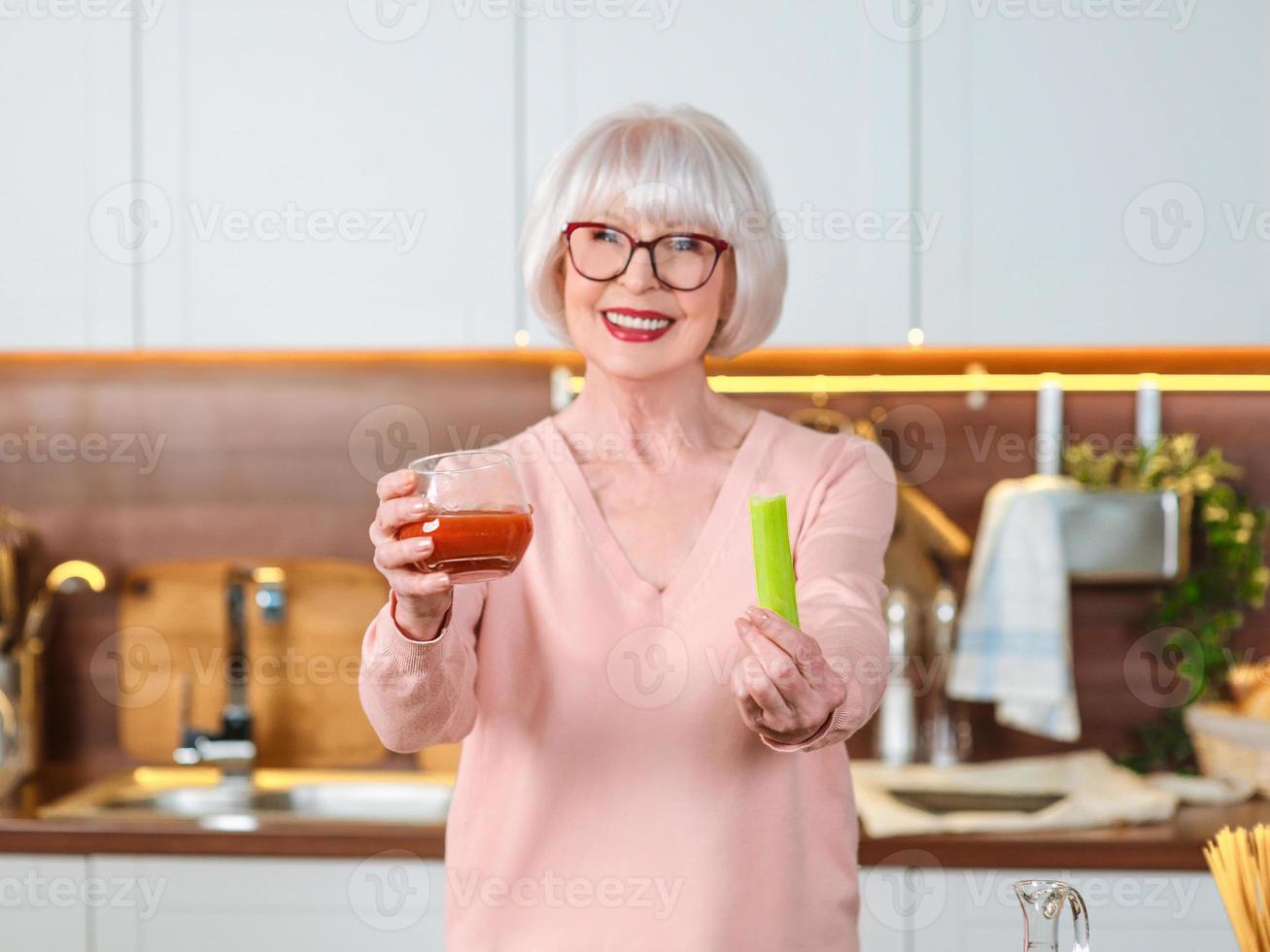 senior donna allegra video blogger che mostra sedano e succo di pomodoro nella sua cucina. crudo, vegetariano, dieta, blogger, concetto di cucina nutrizionista foto