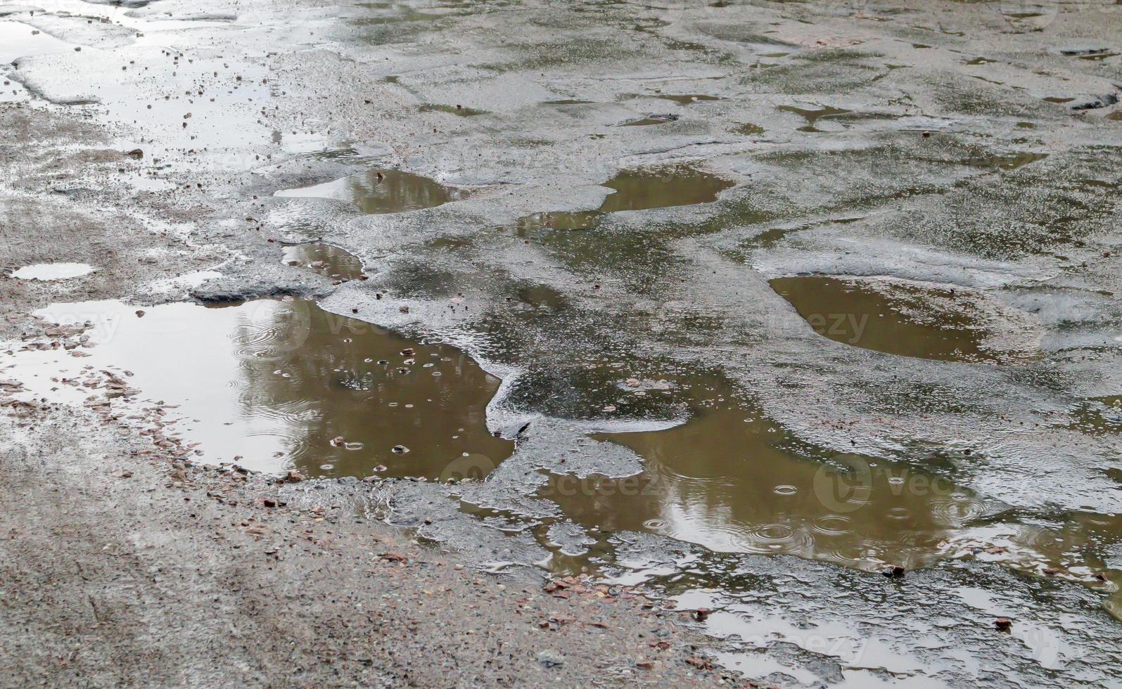 un buco pieno d'acqua in una strada asfaltata. un giorno di pioggia in una grande città, le auto stanno guidando lungo la vecchia strada. strada dissestata con asfalto in fosse e buche pericolose per automobilisti e pedoni. foto