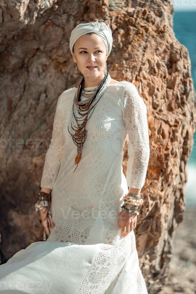 mezzo anziano donna sembra bene con biondo capelli, boho stile nel bianca lungo vestito su il spiaggia decorazioni su sua collo e braccia. foto