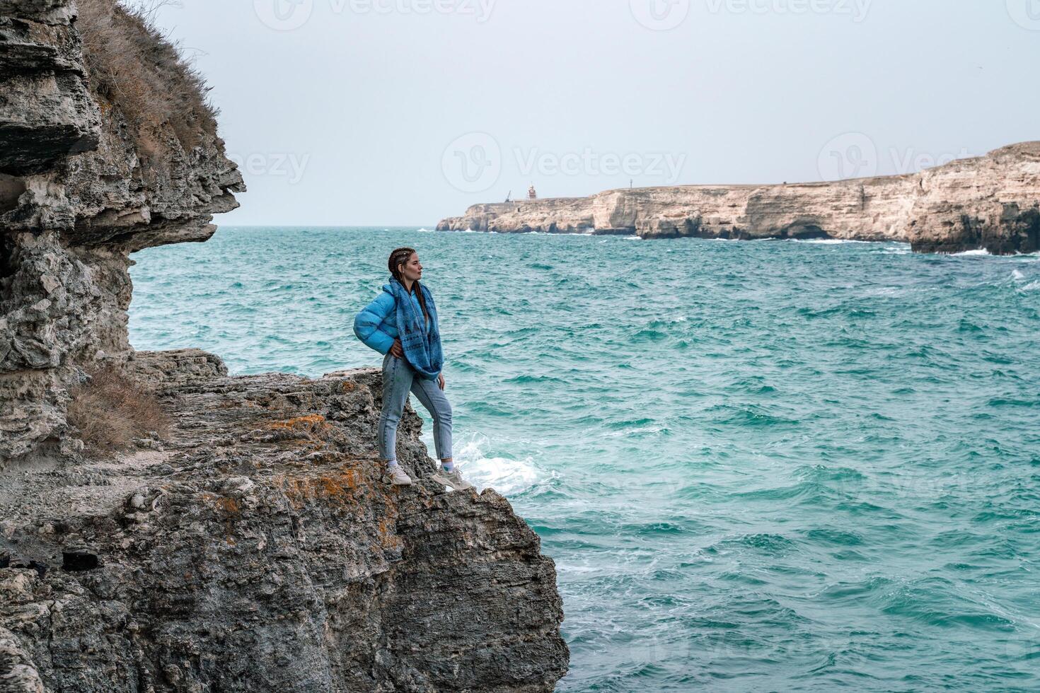 un' donna nel un' blu giacca sta su un' roccia sopra un' scogliera sopra il mare e sembra a il furioso oceano. ragazza viaggiatore riposa, pensa, sogni, gode natura. pace e calma paesaggio, ventoso tempo atmosferico. foto