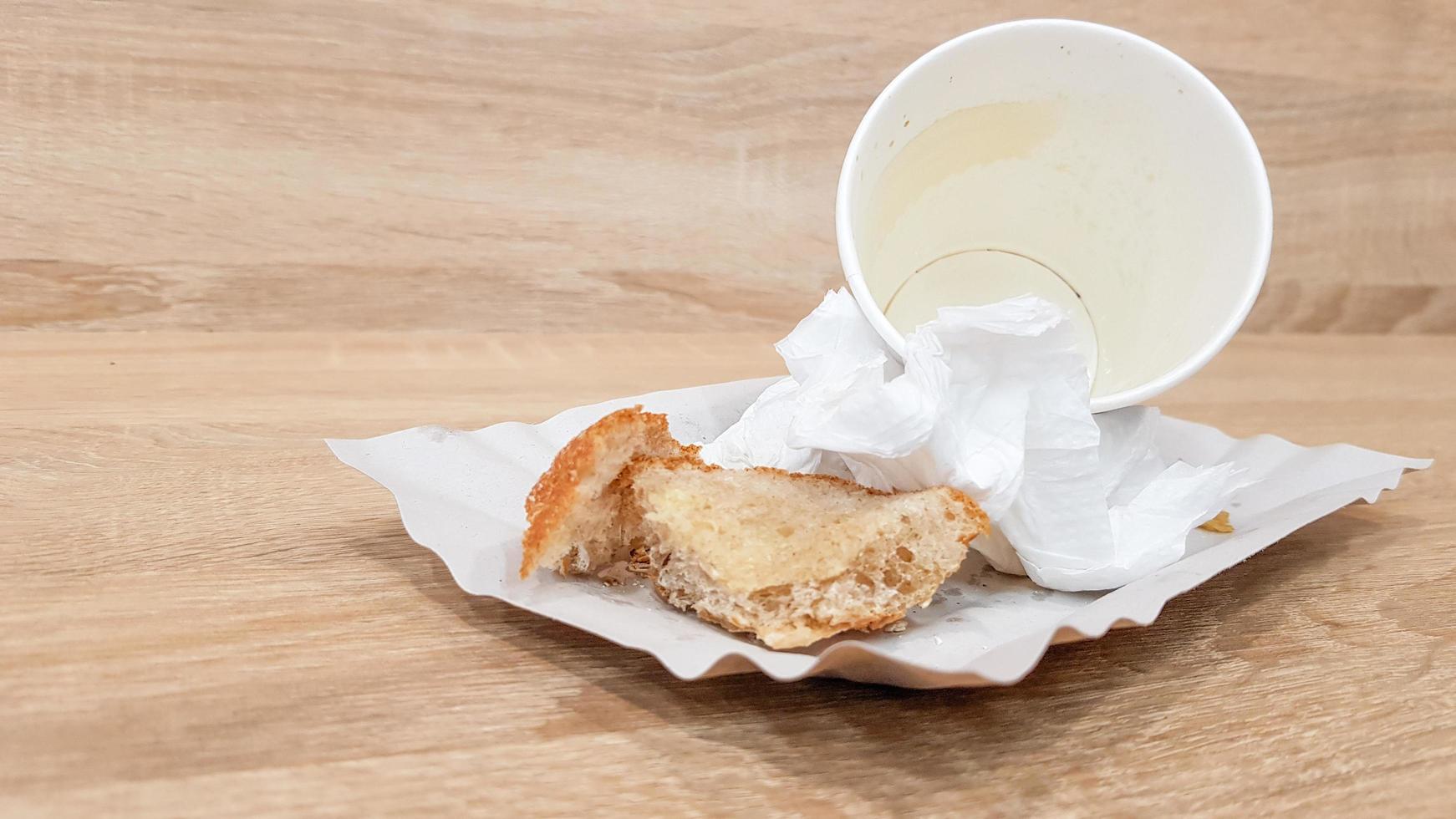avanzi di cibo su un piatto di carta e tazza di caffè di carta sporca vuota su un tavolo di legno in un fast food cafe foto