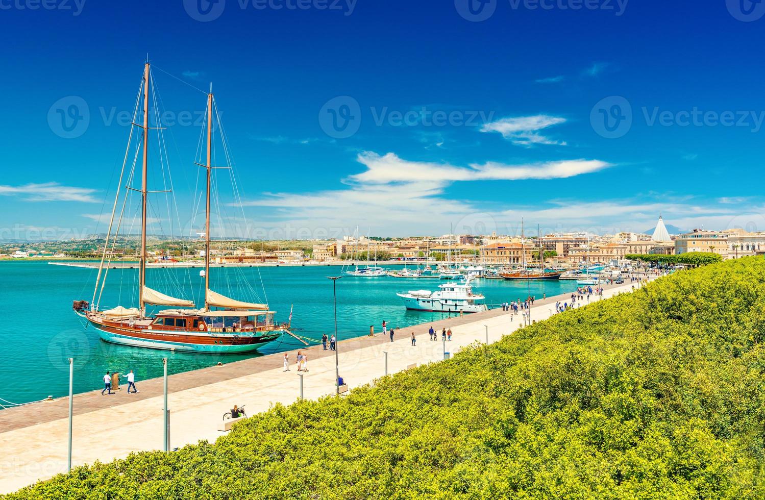 bella vista di una passeggiata a siracusa con gente che cammina. paesaggio urbano dell'isola di ortigia con porto, argine, parco e navi foto