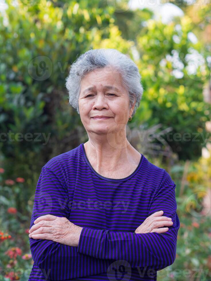 anziano donna con corto bianca capelli in piedi sorridente e braccio attraversato nel il giardino. asiatico anziano donna salutare e avere positivo pensieri su vita rendere sua contento ogni giorno. Salute concetto foto