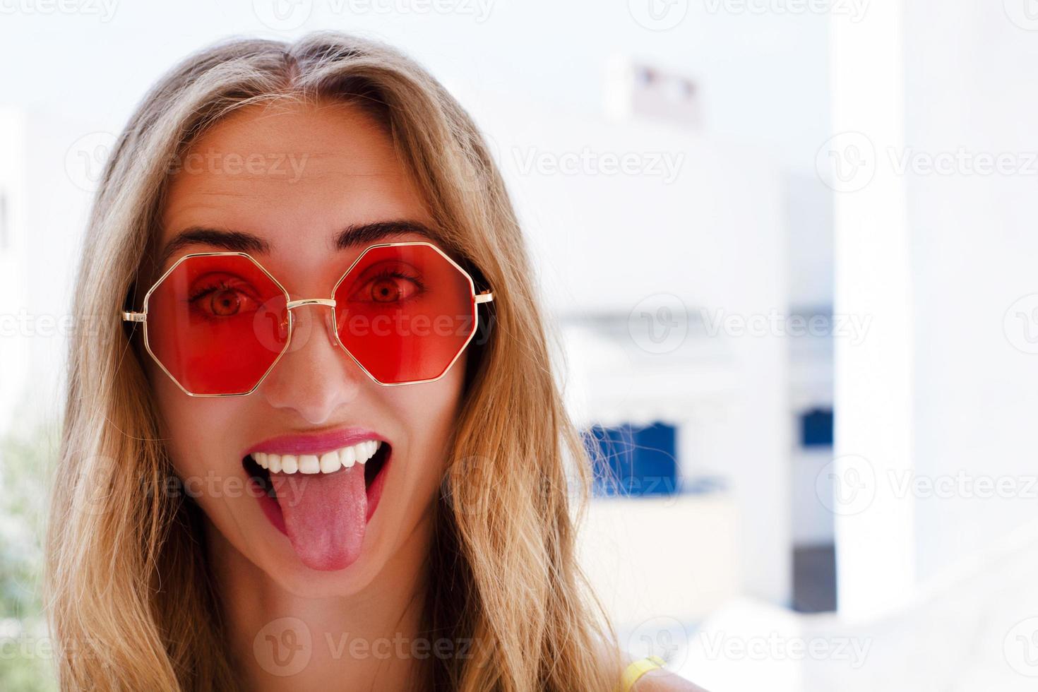 il ritratto di bella giovane donna emotiva in occhiali da sole rossi estivi su uno sfondo di mare mostra la lingua foto