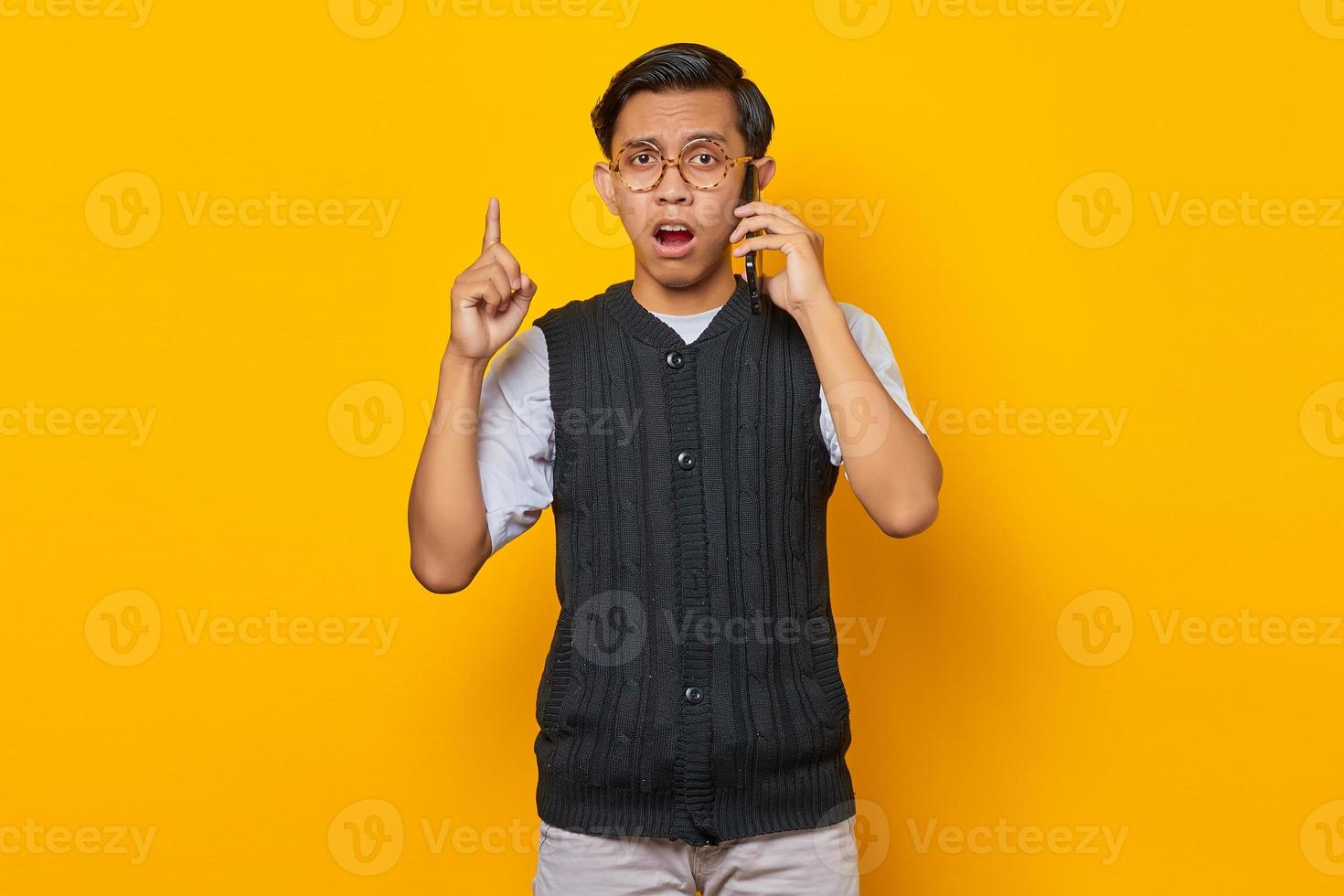 sorpreso giovane uomo asiatico che parla al telefono cellulare e punta il dito su sfondo giallo foto