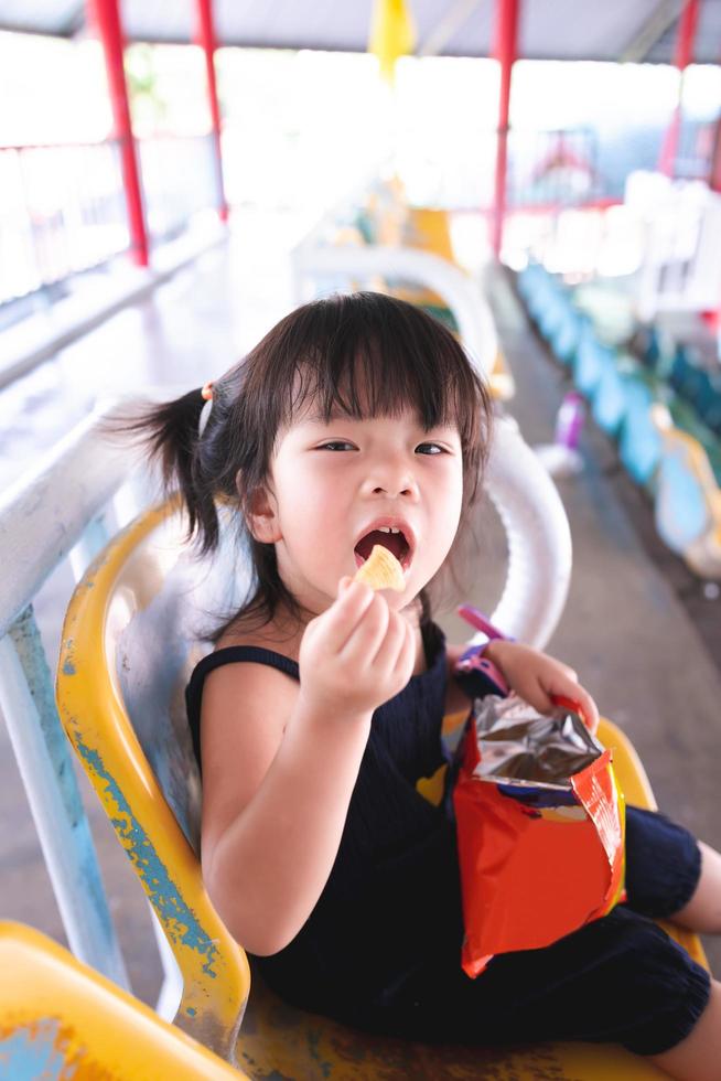 felice ragazza carina di 3 anni che mangia snack in tribuna. bambino raccoglie cibo croccante in bocca. immagine verticale. foto