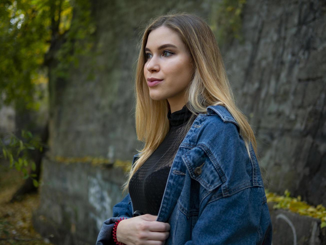 il ritratto di un'attraente ragazza bionda con una giacca di jeans è in piedi per strada contro un muro di pietra foto