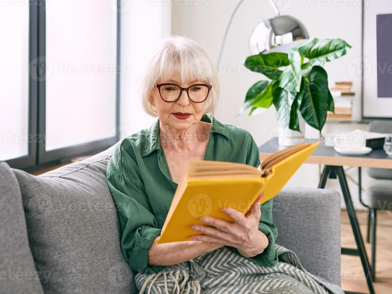 allegra donna anziana seduta sul divano a leggere un libro a casa. educazione, maturo, concetto di svago foto
