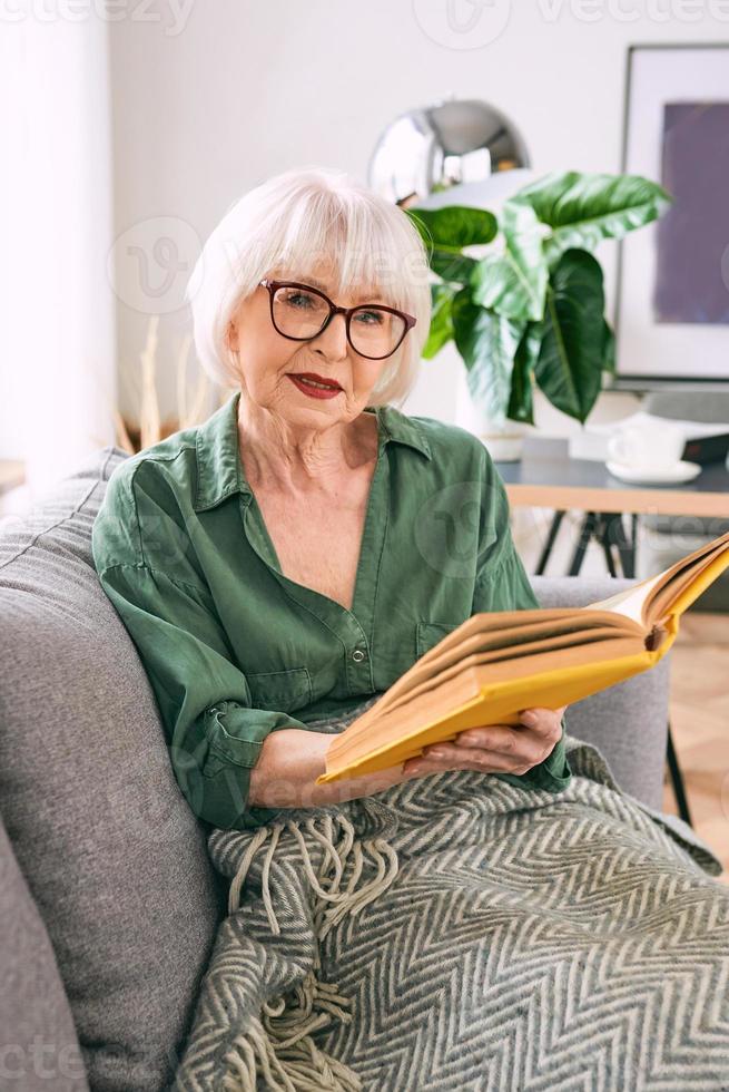allegra donna anziana seduta sul divano a leggere un libro a casa. educazione, maturo, concetto di svago foto
