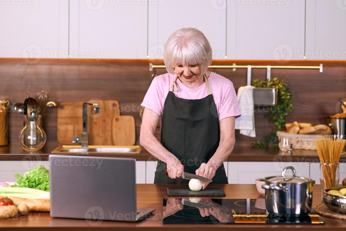 la donna allegra senior sta cucinando alla cucina moderna dal computer portatile. cibo, educazione, concetto di stile di vita foto