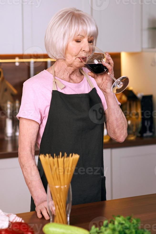 donna allegra senior sta bevendo vino rosso durante la cottura in cucina moderna. cibo, educazione, concetto di stile di vita foto