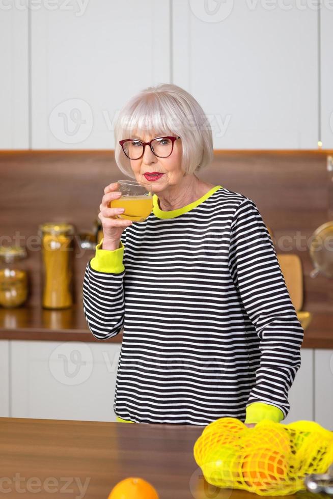 allegra donna sorridente piuttosto anziana in maglione a righe che beve succo d'arancia mentre si trova in cucina. stile di vita sano e succoso, casa, concetto di persone anziane. foto