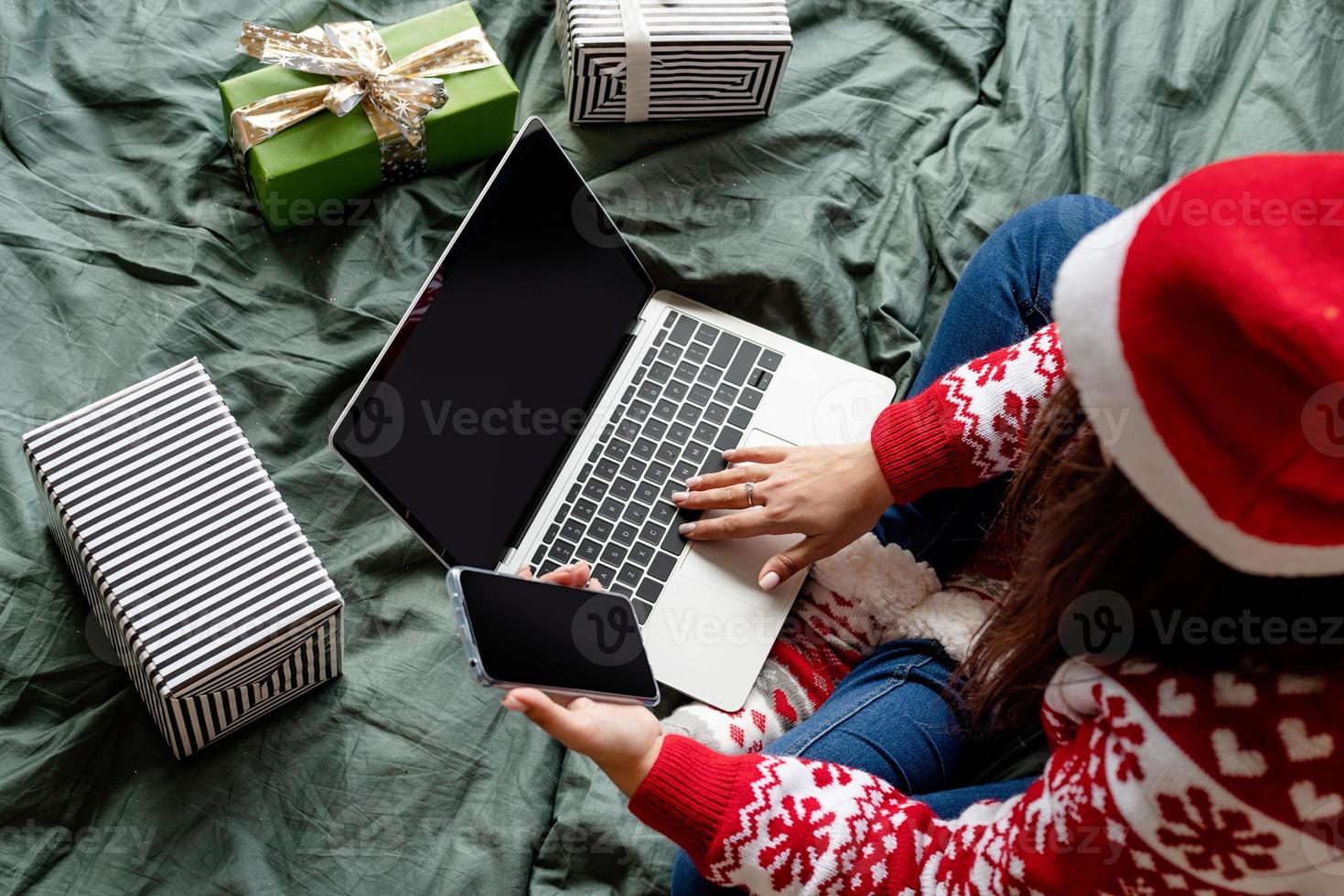 donna che indossa abiti natalizi seduta sul letto usando laptop e cellulare foto