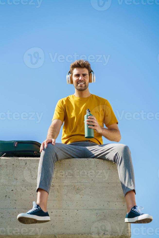 maschio seduto fuori con una bottiglia d'acqua in alluminio, cuffie e zaino. foto