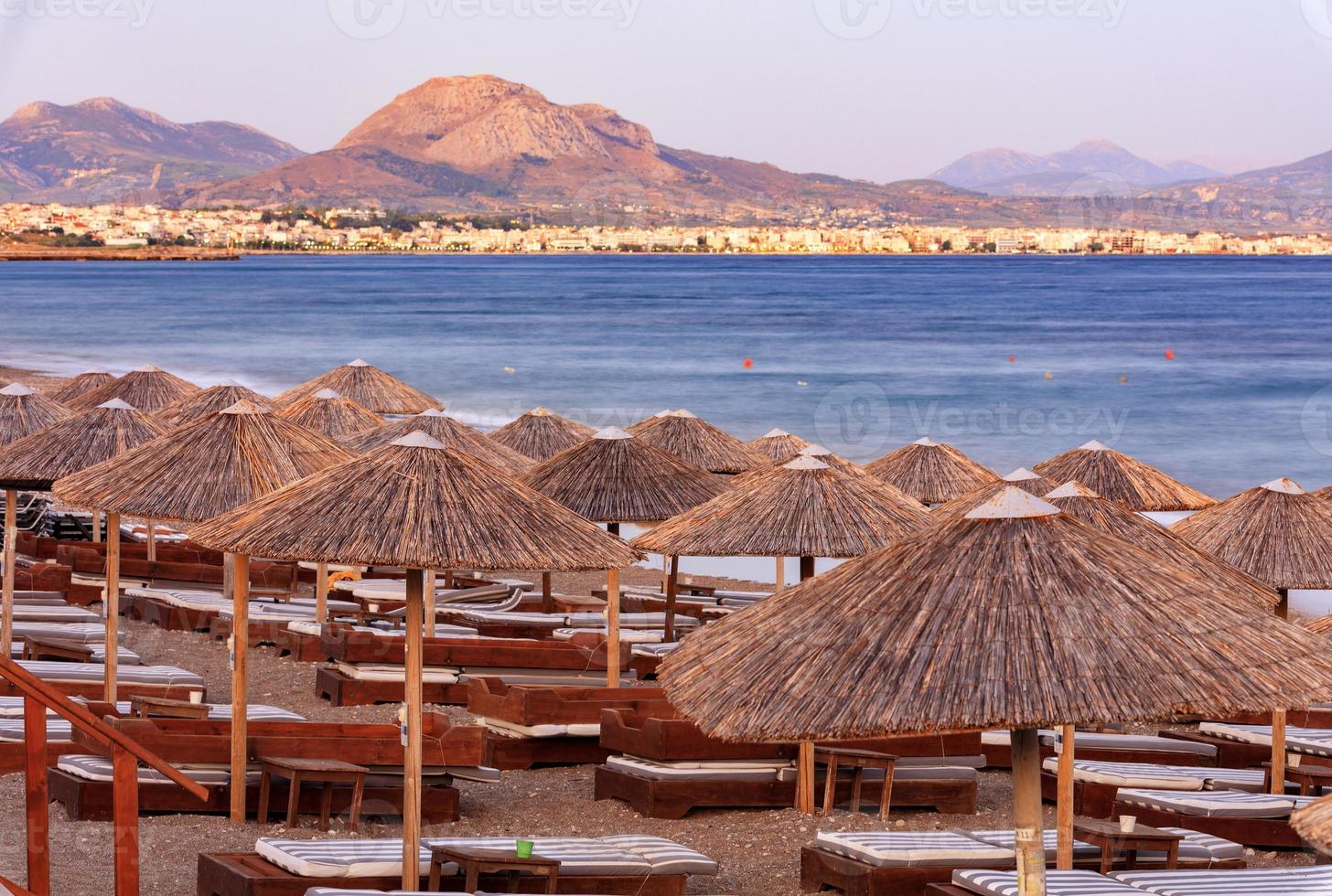 le cime di paglia degli ombrelloni e delle sedie a sdraio in legno con i materassini sul lungomare deserto sotto i raggi del sole al tramonto della sera. foto