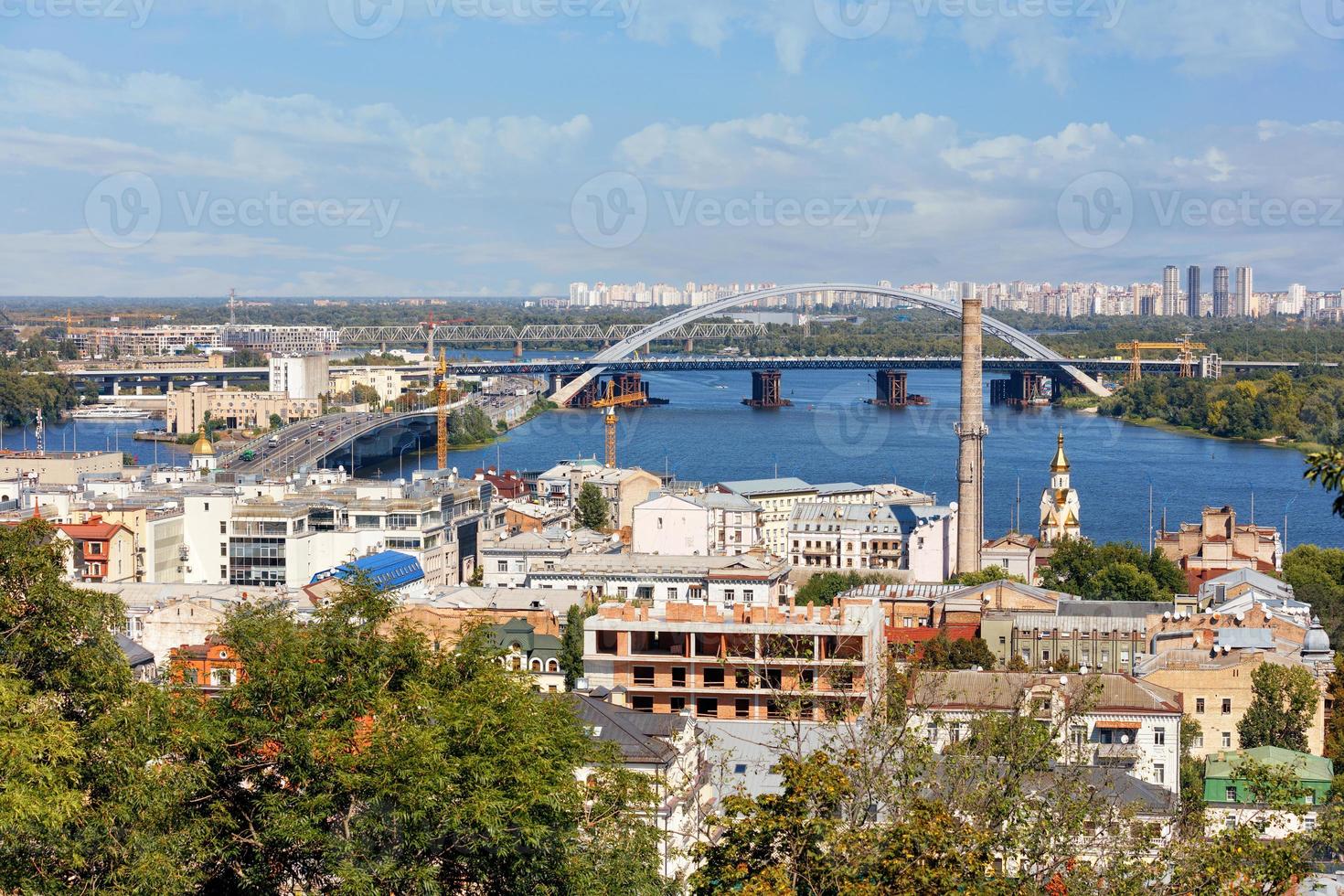 il paesaggio dell'estate kyiv con vista sul vecchio quartiere di podil con ponti stradali e ferroviari, un camino di un vecchio locale caldaia e un campanile con cupola dorata, il fiume dnipro. foto