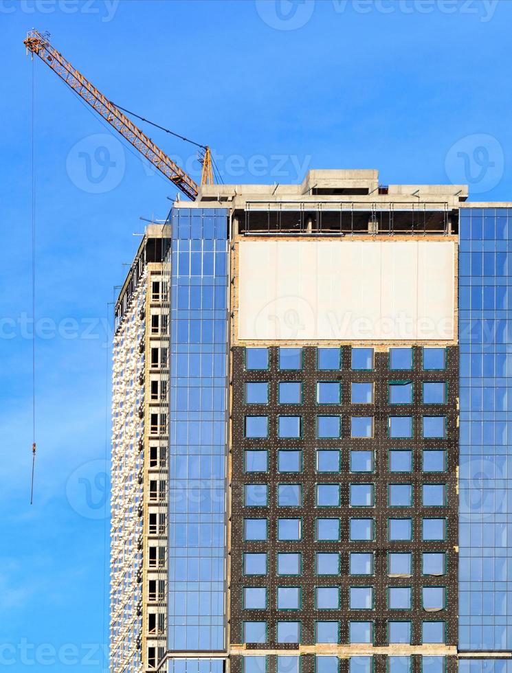 costruzione di un moderno edificio in cemento con facciata in vetro, riflesso del cielo azzurro e gru a torre. foto