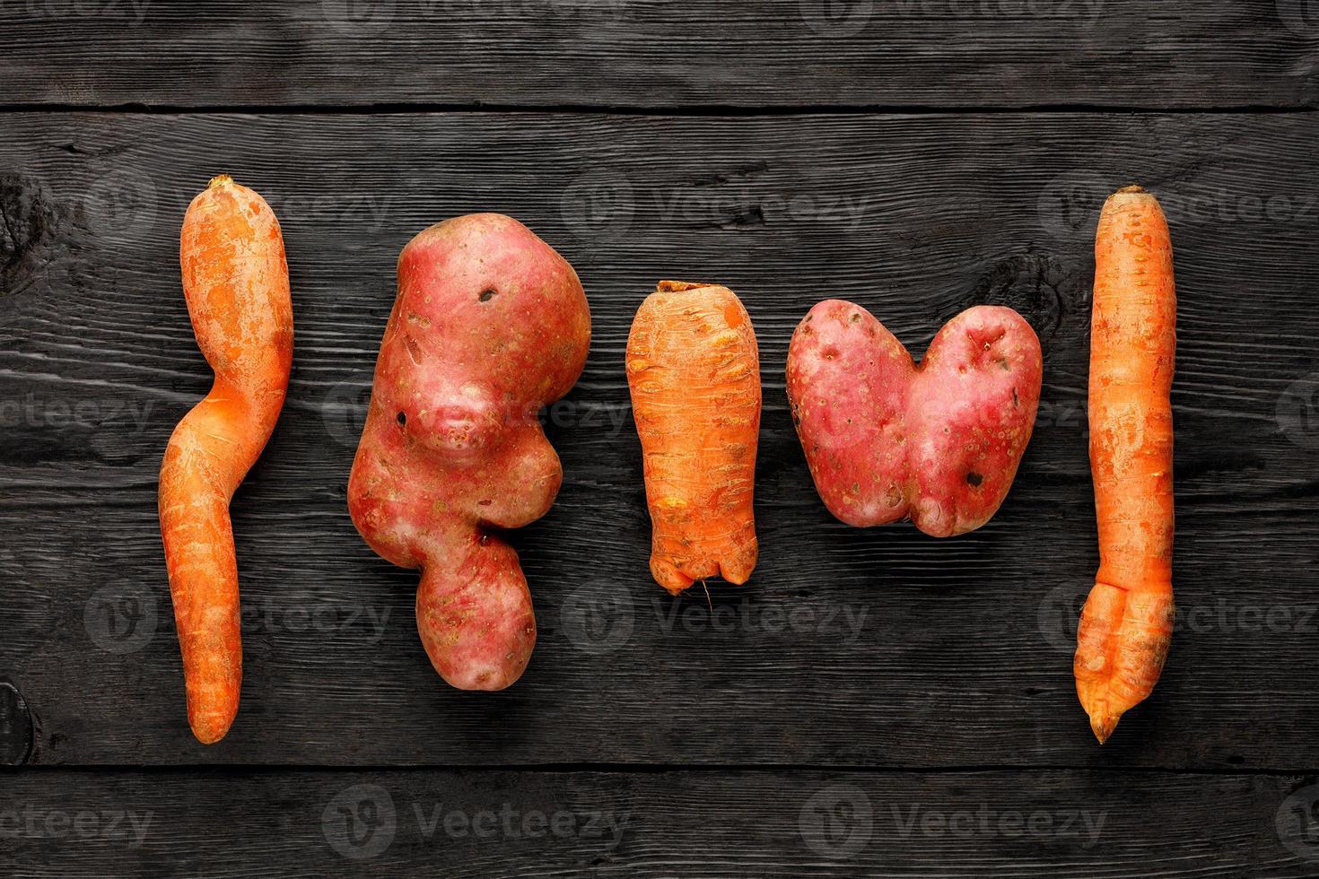 patata brutta a forma di cuore e carota contorta su fondo di legno nero. concetto di rifiuti vegetali o alimentari. vista dall'alto, primo piano. foto