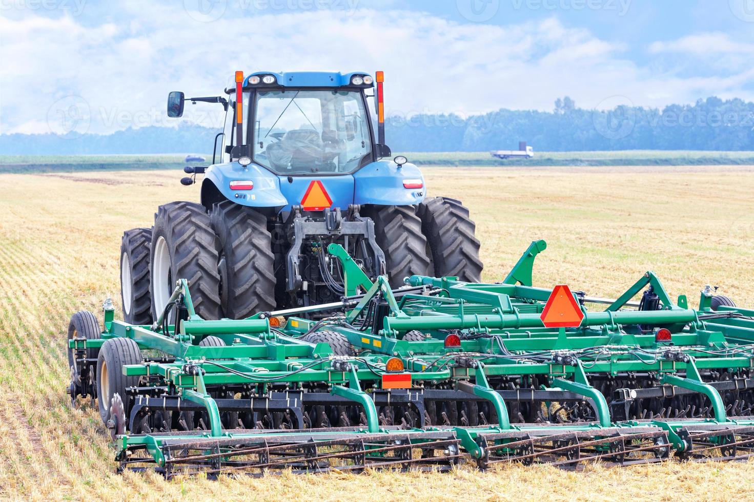 un trattore agricolo con un intoppo, un erpice, si erge sullo sfondo di un campo di grano raccolto. foto