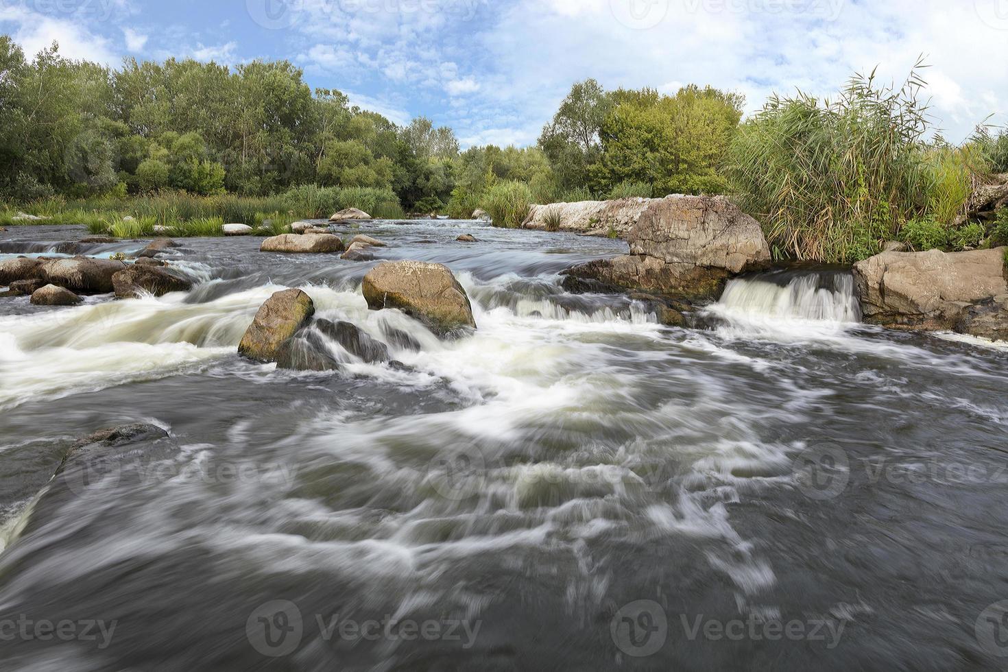 il rapido flusso del fiume, le coste rocciose, le rapide, la vegetazione verde brillante e il cielo azzurro e nuvoloso in estate - una vista frontale foto