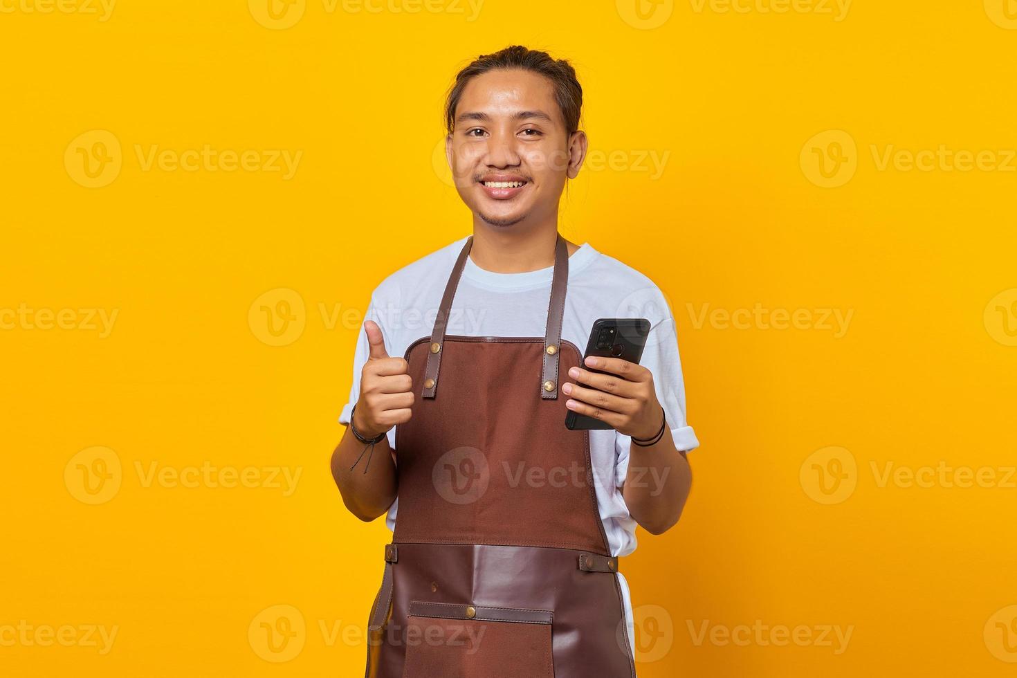 uomo asiatico eccitato e allegro che tiene smartphone e pollice in alto su sfondo giallo foto