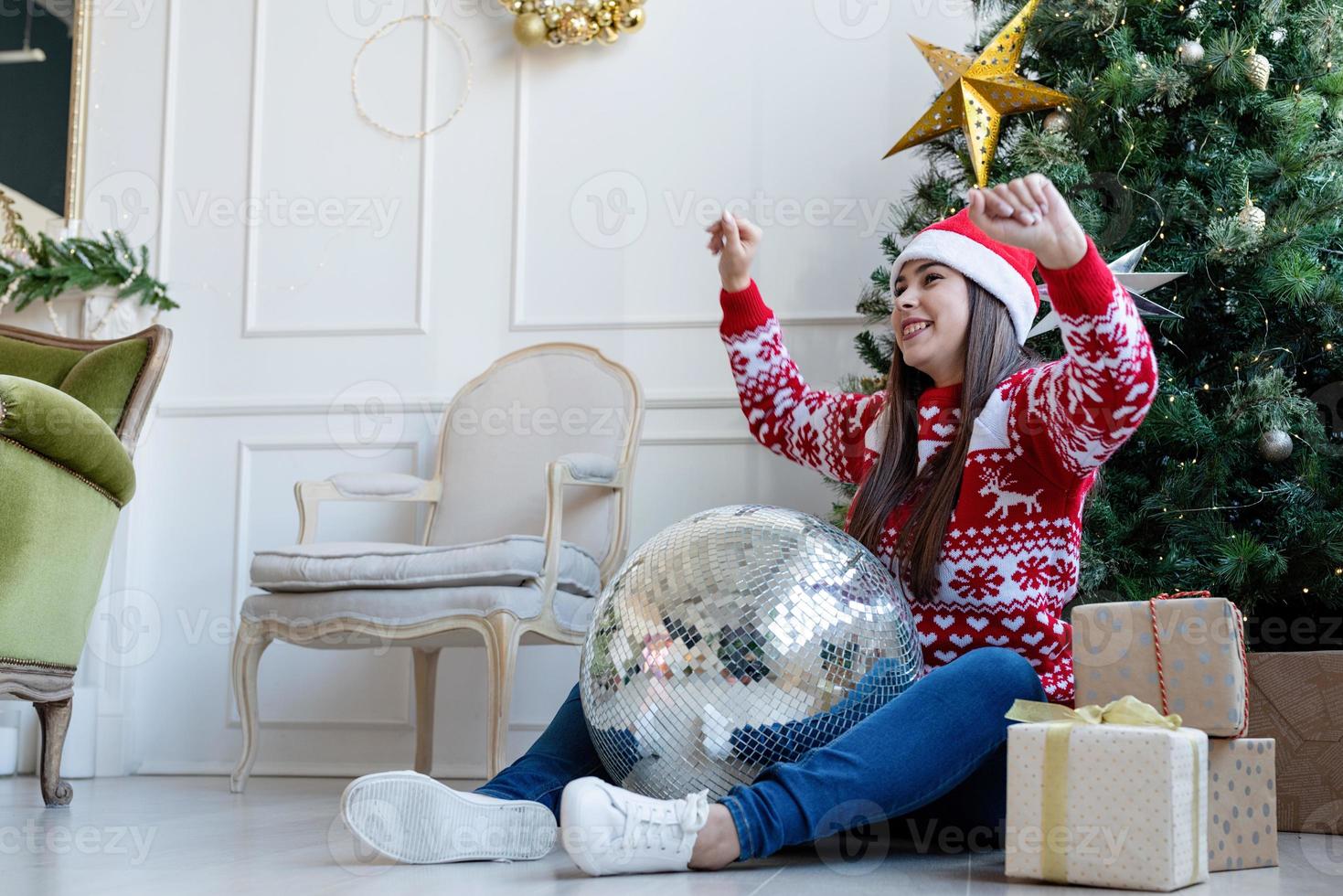 giovane donna con cappello da Babbo Natale che tiene in mano una palla da discoteca a specchio che balla vicino all'albero di natale foto