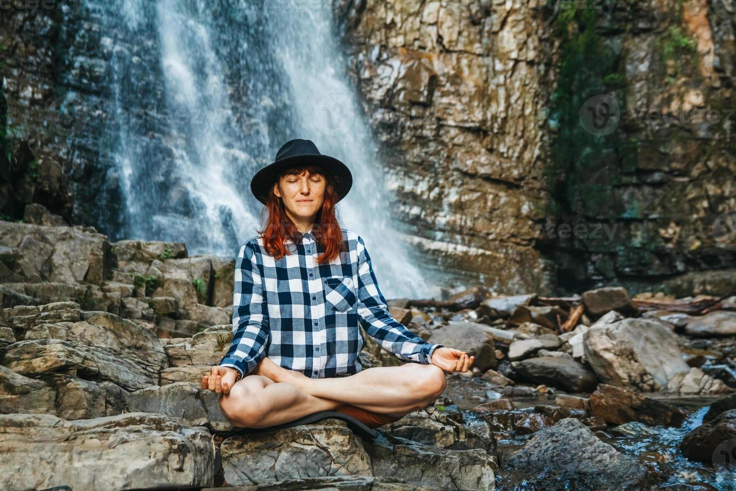 donna che medita su rocce nella posizione del loto contro una cascata foto