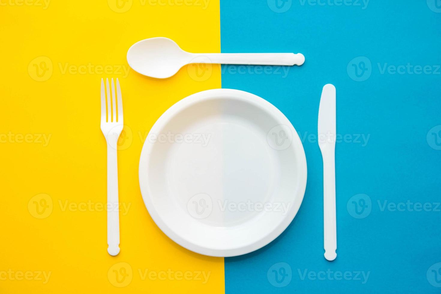forchetta, coltello, cucchiaio e piatto di plastica bianchi su sfondo giallo e blu foto