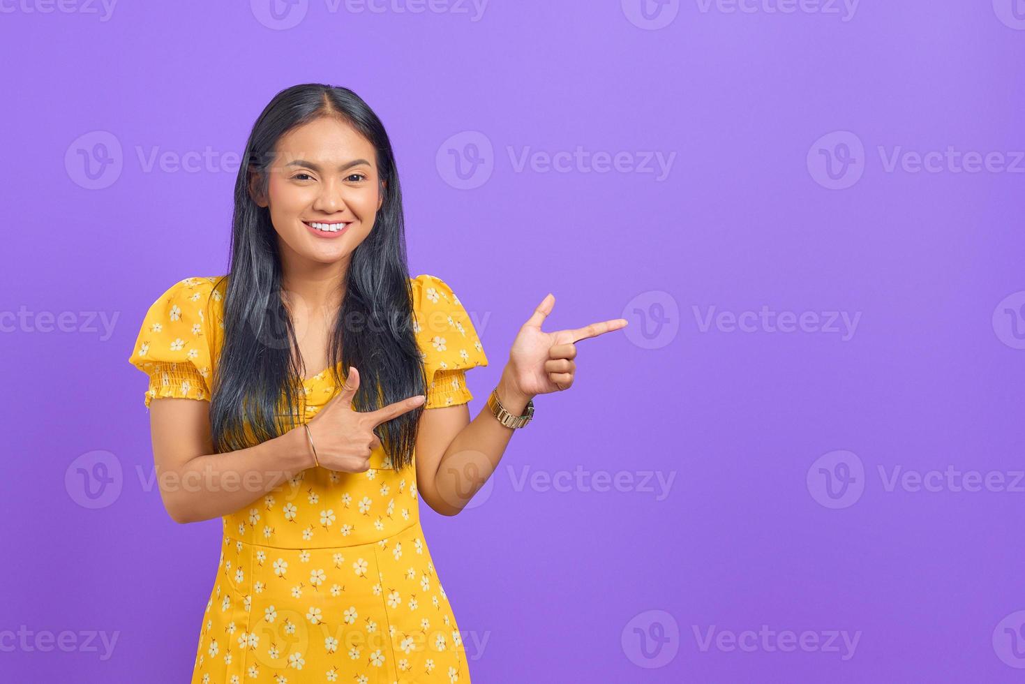 ritratto di giovane donna asiatica sorridente che punta il dito contro lo spazio della copia su sfondo viola foto