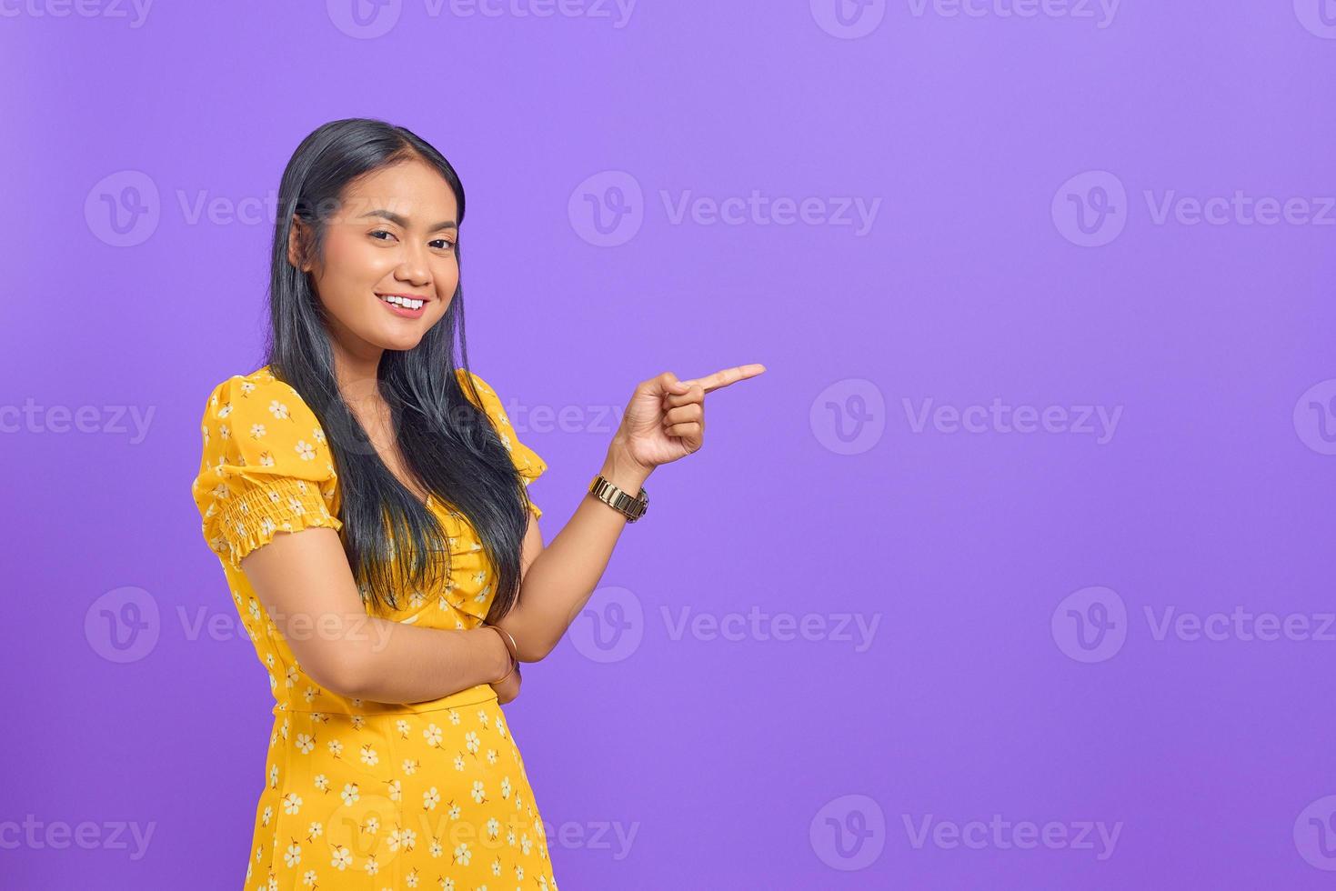 ritratto di giovane donna asiatica sorridente che punta il dito contro lo spazio della copia su sfondo viola foto