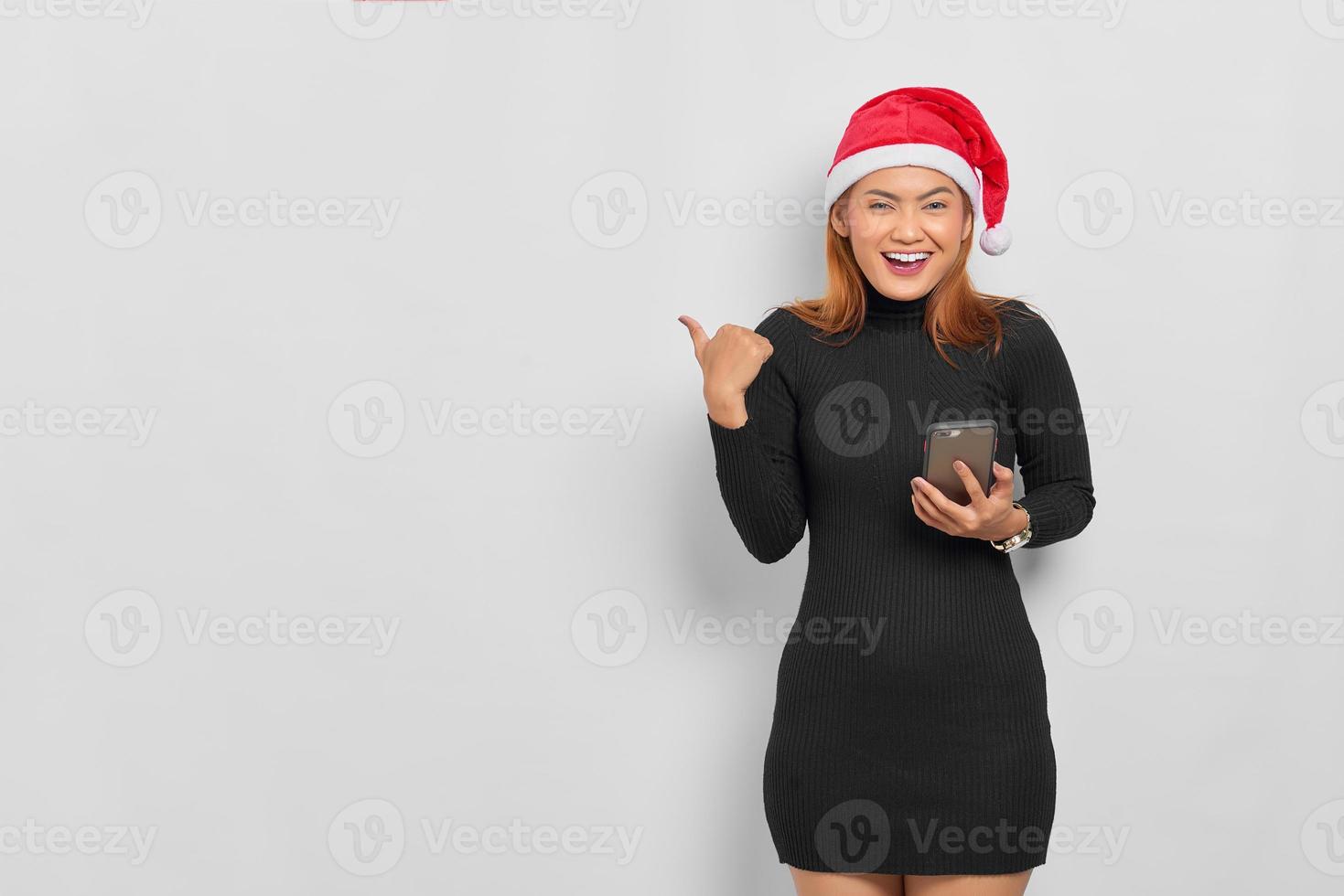 Sorridente giovane donna asiatica in santa claus hat tenendo il telefono cellulare e puntare il dito contro lo spazio della copia isolato su sfondo bianco foto