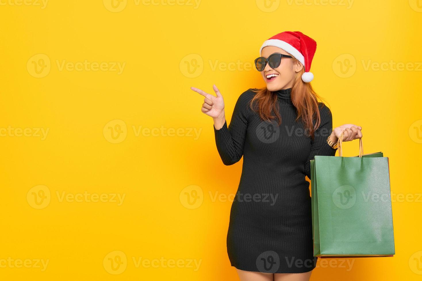 Allegra giovane donna asiatica con cappello di Babbo Natale e occhiali da sole che tengono le borse della spesa, puntando il dito contro lo spazio della copia isolato su sfondo giallo foto