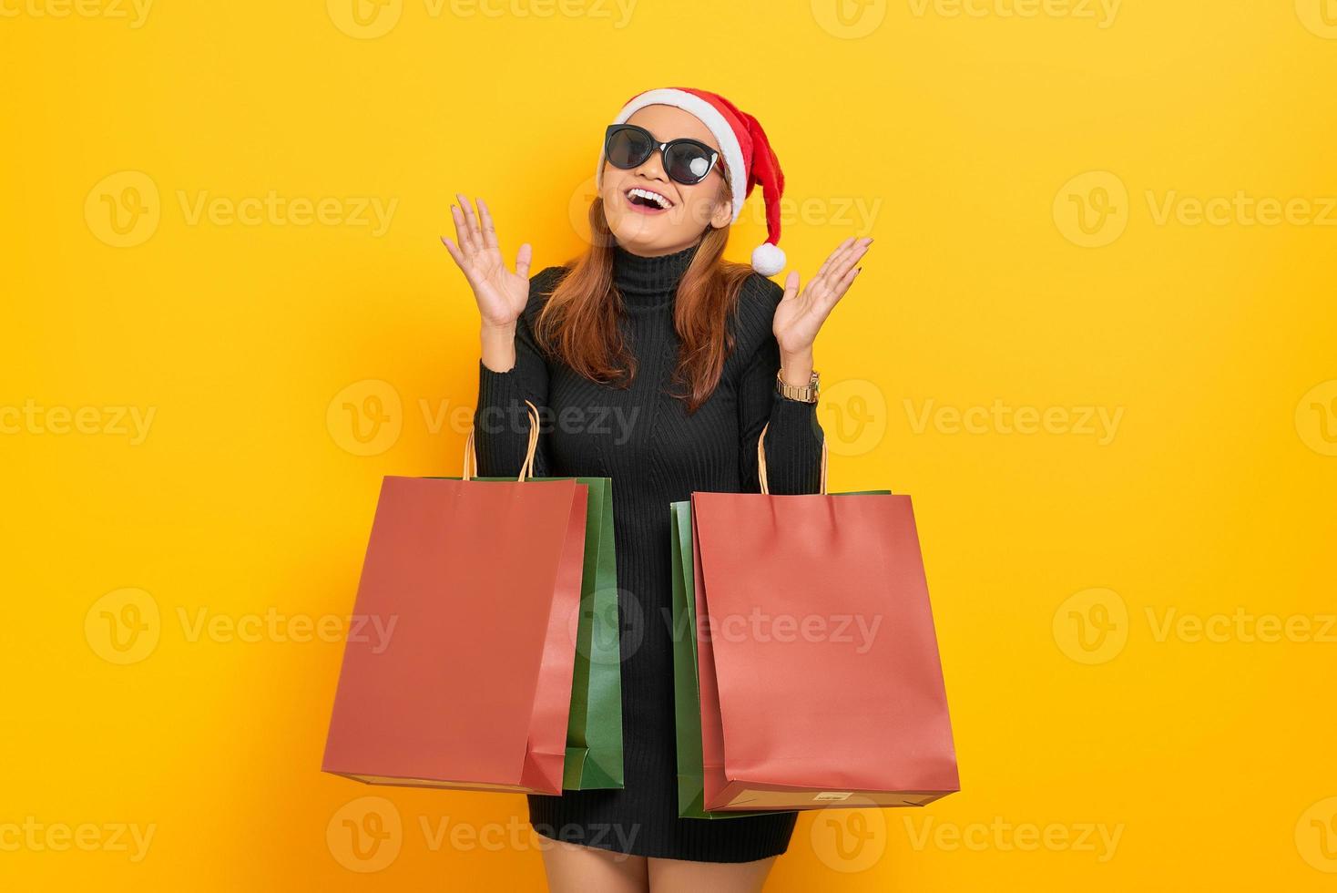Allegra giovane donna asiatica in cappello di Babbo Natale e occhiali da sole che tengono le borse della spesa e celebrano la fortuna isolate su sfondo giallo foto