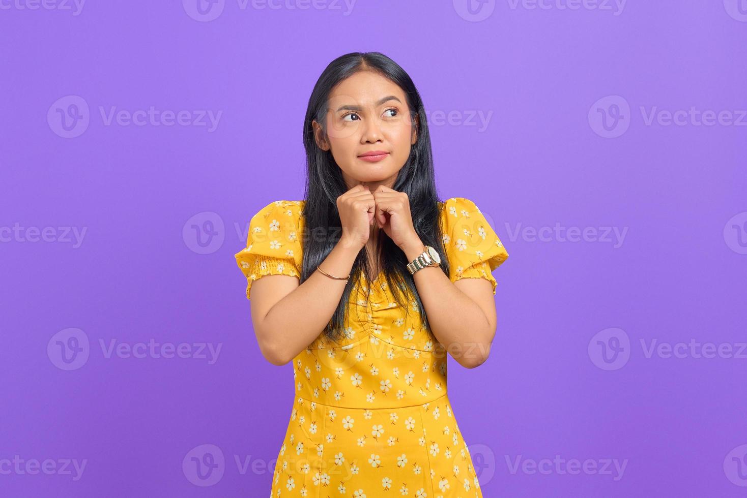 Attraente giovane donna asiatica tiene la mano sotto il mento e guarda da parte isolata su sfondo viola foto
