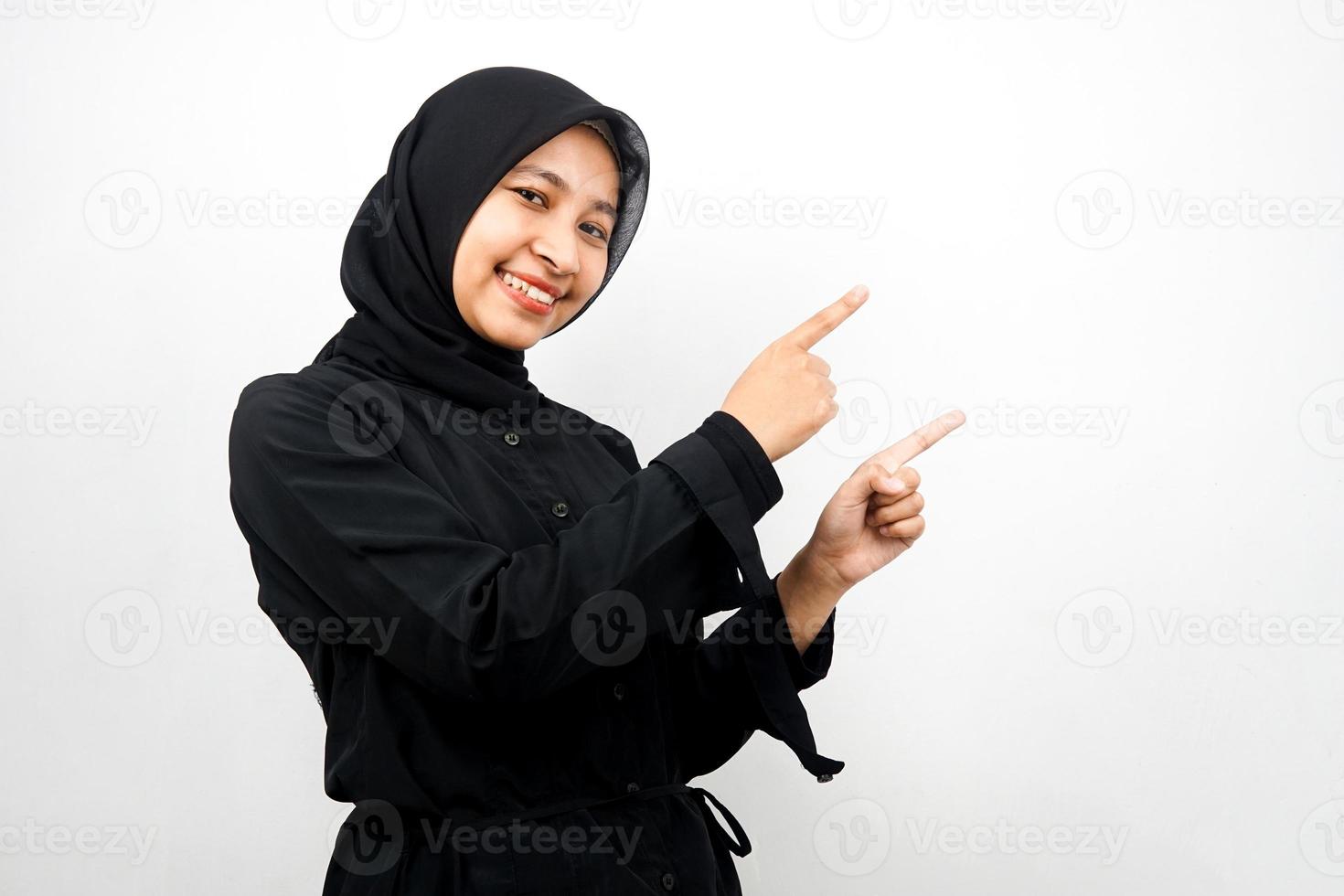 bella giovane donna musulmana asiatica sorridente sicura di sé, entusiasta e allegra con le mani che puntano lo spazio vuoto presentando qualcosa di fronte alla telecamera isolata su sfondo bianco, concetto di pubblicità foto