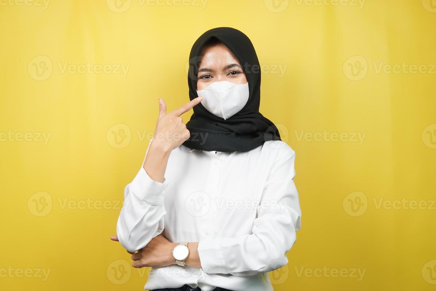donna musulmana che indossa una maschera bianca, con la mano che punta alla maschera, previene il gesto del virus corona, previene il covid-19, isolato su sfondo giallo foto