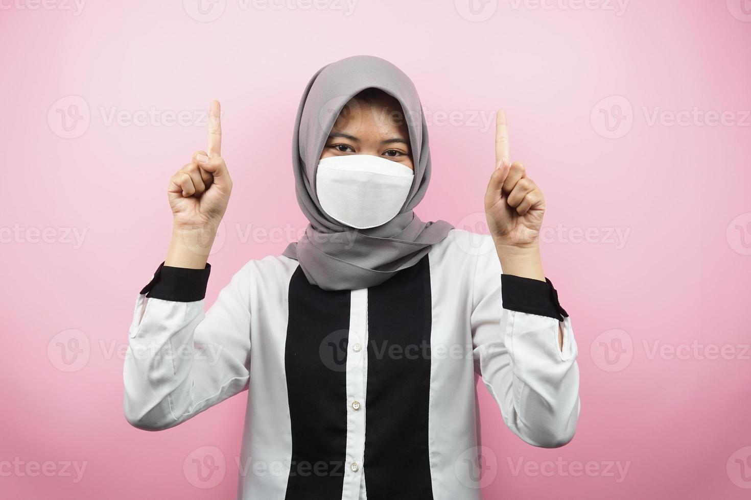 donna musulmana che indossa una maschera bianca, mano che punta allo spazio vuoto, mano che punta verso l'alto presentando qualcosa, isolata su sfondo rosa foto