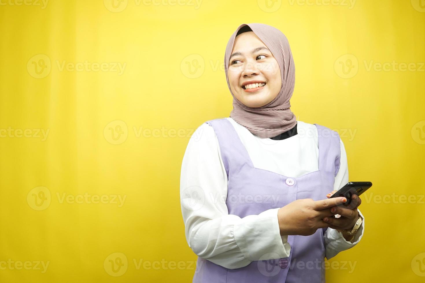 bella giovane donna musulmana asiatica sorridente sicura di sé, entusiasta e allegra con le mani che tengono lo smartphone, vista guardando lo spazio vuoto che presenta qualcosa, isolato su sfondo giallo foto