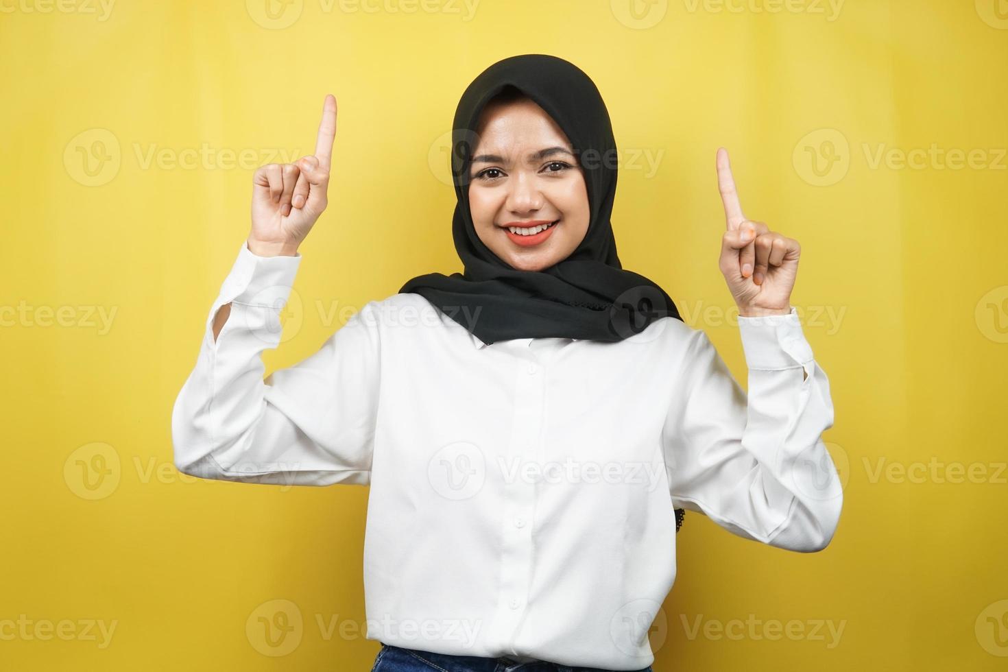 bella giovane donna musulmana asiatica sorridente sicura di sé, entusiasta e allegra con le mani rivolte verso l'alto presentando qualcosa, guardando la telecamera isolata su sfondo giallo, concetto pubblicitario foto