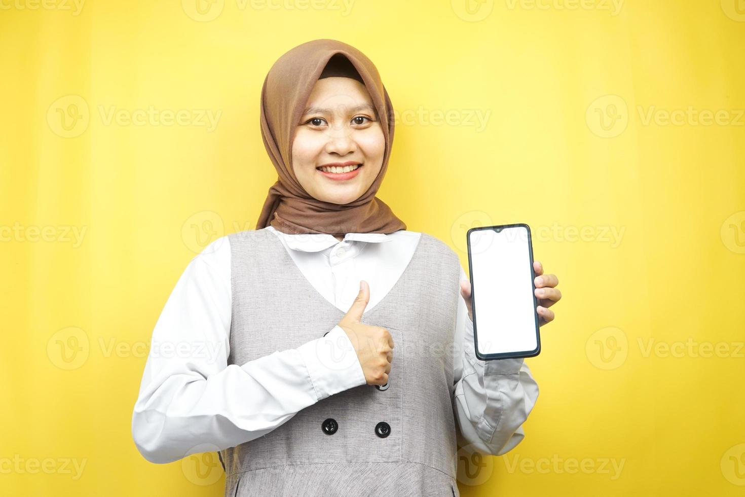 bella giovane donna musulmana asiatica sorridente sicura di sé, entusiasta e allegra con la mano che tiene lo smartphone, promuovendo qualcosa, app per la promozione della mano, isolata su sfondo giallo foto