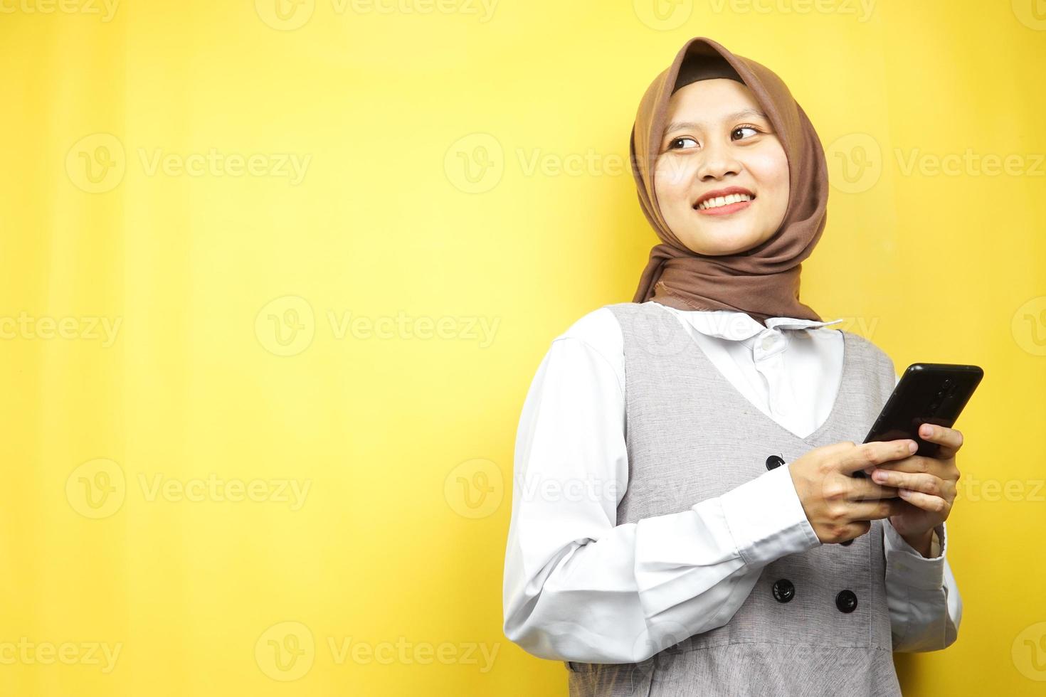 bella giovane donna musulmana asiatica sorridente sicura di sé, entusiasta e allegra con le mani che tengono lo smartphone, vista guardando lo spazio vuoto che presenta qualcosa, isolato su sfondo giallo foto